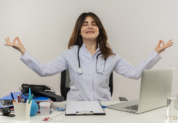 with closed eyes smiling middle-aged female doctor wearing wearing medical robe with stethoscope sitting at desk work on laptop with medical tools showing okey gesture on isolated white backgroung with copy space